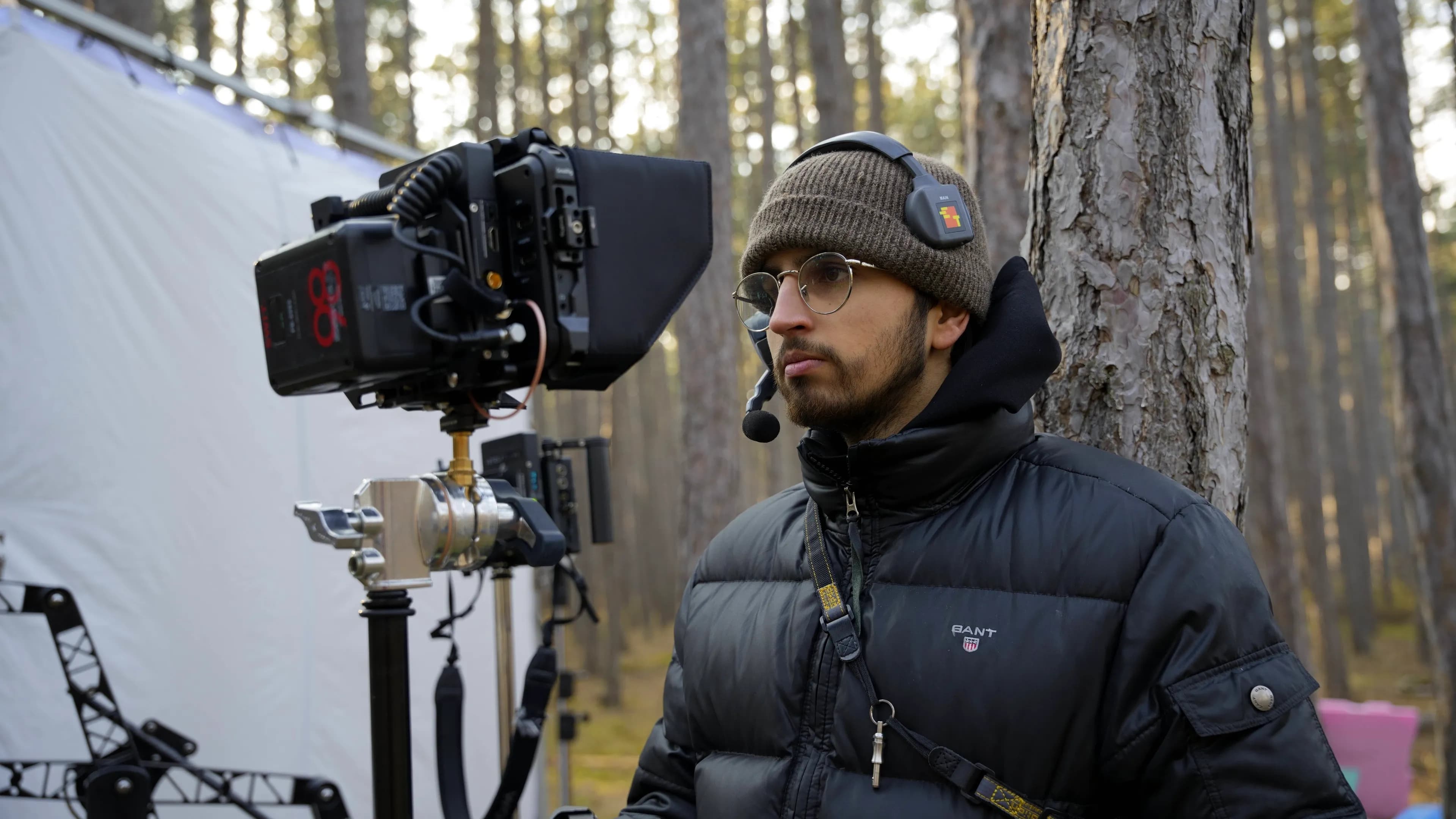 Will Rose, is captured in a medium shot outdoors. He is looking intently towards the right, wearing a dark, quilted puffer jacket, a textured knitted beanie, and clear-framed round glasses. A communications headset with a boom microphone is over his ears. To his left, a professional digital cinema camera rig with a large matte box is prominently displayed on a tripod. The background is a softly blurred forest of tall, slender trees, with a large white fabric surface or screen visible on the far left, suggesting a film set.